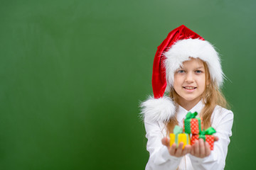 Smiling girl wearing a red christmas hat holds gift boxes near green chalkboard. Empty space for text