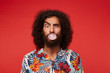 Indoor shot of funny brunette curly male with beard inflating ball with chewing gum while looking at camera with raised eyebrow, isolated against red background in casual clothes