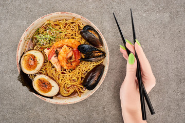 Wall Mural - cropped view of woman holding chopsticks near spicy seafood ramen on grey surface