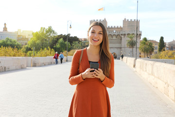 Wall Mural - Portrait of happy young tourist woman walking in Valencia city holding smart phone looking at camera.