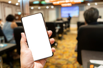 Hand holding the smartphone with blank screen on blurred image of people in conference room for meeting seminar as background. White screen in smart phone with clipping path