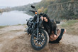 Gorgeous brunette girl with long hair in a black leather jacket sits near a modern motorcycle on a background of nature. Portrait of a sexy woman near an expensive black bike.