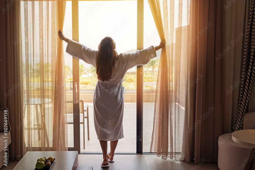 Young woman wearing white bathrobe opening curtains in luxury hotel room - obrazy, fototapety, plakaty 
