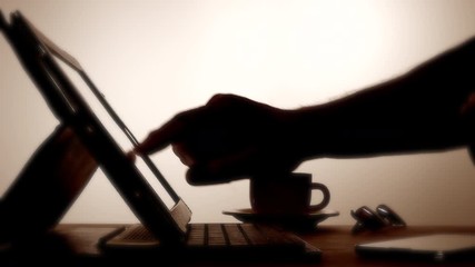Backlit dolly shot of a man’s hands using a keyboard and swiping a tablet computer.