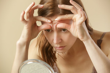 Portrait of a young brunette woman, in her thirties, examining her face in front of mirror, aging, fine wrinkles and skin complexion concept