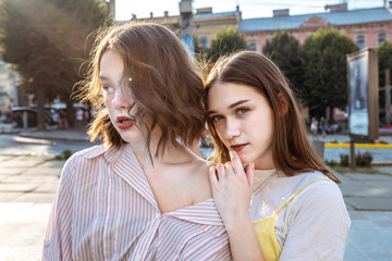 Wall Mural - Young sensual brunette girls in casual clothes standing together among the street, girl in white t-shirt holds her hand on the shoulder of other girl with wavy hair and freckles