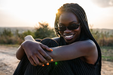 Close Up Portrait Of The Young Happy African Girl In Sunglasses That Sits On The Dirt Road, Smiling And Looking At The Camera