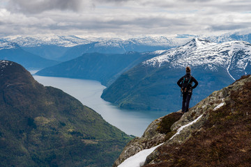 An explorer stands atop a mountain looking across a sweeping view of Norwegian fjords and snow capped mountains