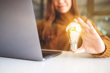 Wall Mural - Closeup image of a businesswoman holding and showing a glowing light bulb while working on laptop