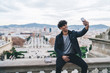 © BullRun - Happy cheerful Latino male traveller dressed in stylish outfit resting during Spanish vacations to Barcelona city clicking selfie pictures on Montjuic area, millennial man photographing himself