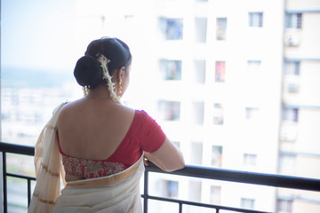 An young and attractive Indian woman in white traditional sari and red blouse and flowers is turning back while standing in a balcony for the celebration of Onam/Pongal. Indian lifestyle.