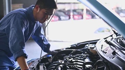 Wall Mural - Young Asian auto mechanic examining engine at car hood in auto repair workshop. Repairman checking for customer to safety maintenance support before long trip. Business and transportation concept.