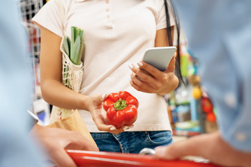 Wall Mural - Daily Shopping. Couple in the supermarket with cart together holding bell pepper browsing smartphone close-up