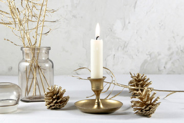 burning candle in vintage metal candlestick, golden cones and grass on white table. golden christmas