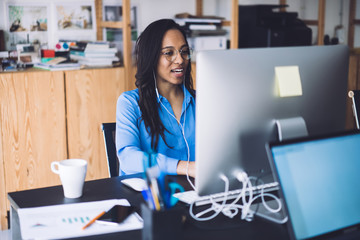 Wall Mural - Young African American woman speaking at work place using earphones and computer