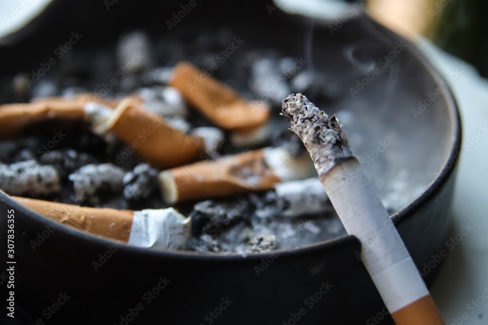 one cigarette in the ashtray one burning cigarette placed on ashtray full of smoked cigarettes, extreme  close up of burning cigarette with blurred ashtray full of cigarette butts,  focus selective Stock Photo