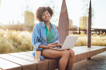 Happy smiling dark skinned woman freelancer satisfied with earning money online checking income on mobile app, prosperous female blogger sitting with laptop computer looking at camera in park