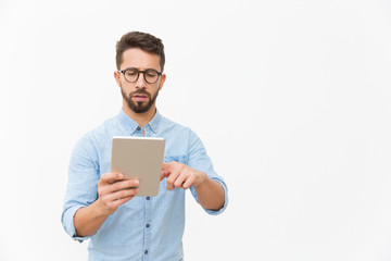 Focused professional working on tablet. Handsome young man in casual shirt and glasses standing isolated over white background. Digital communication concept