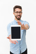 © Mangostar - Positive smiling tablet user showing blank screen at camera. Handsome young man in casual shirt and glasses standing isolated over white background. Communication and advertising concept