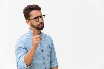 Frowning concerned guy shaking finger at camera. Handsome young man in casual shirt and glasses standing isolated over white background. Warning gesture concept