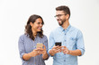 © Mangostar - Happy cheerful couple with cellphones chatting and laughing. Young woman in casual and man in glasses posing isolated over white background. Good news concept