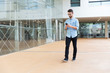 © Mangostar - Positive guy with smartphone texting message while going through hotel lobby. Handsome young man in casual shirt and glasses walking indoors with glass wall in background. Communication concept