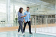 Joyful colleagues in casual looking at cellphone screen together while going through office hallway. Young man and woman walking indoors with glass wall in background. Communication concept