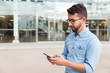 © Mangostar - Focused guy in eyewear texting message or consulting internet on smartphone. Handsome young man in casual shirt and glasses standing at outdoor glass wall. Digital communication concept
