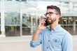 © Mangostar - Pensive guy in eyewear speaking on cellphone and looking into distance. Handsome young man in casual shirt and glasses standing at outdoor glass wall. Phone talk concept