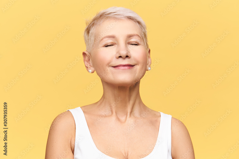 Fashionable beautiful retired Caucasian woman with pixie hairdo wearing casual clothes posing in studio keeping eyes closed and smiling with pleasure and enjoyment, listening to good music or dreaming - obrazy, fototapety, plakaty 