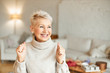 Indoor shot of fashionable overjoyed mature female in turtleneck sweater enjoying positive news, having ecstatic facial expression, laughing and clenching fists. Success and achievements concept
