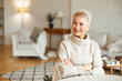 People, lifestyle and mature age concept. Indoor shot of middle aged short haired woman sitting in her stylish cozy living room crossing arms on chest and looking away with smile, wearing sweater