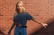 © Vasya - Attractive young blonde girl with red lips dressed in grey striped t-shirt withopen hands looking at the camera isolated over red brick wall