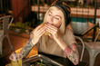 Horizontal photo of pretty young blonde woman with tattooes posing over city cafe interior, enjoying taste of hamburger with closed eyes, wearing trendy clothes