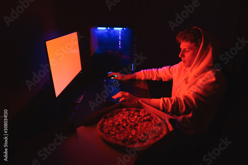Cuadros en Lienzo Young man in a hoodie sits at work at a table with a pizza delivery box and uses a computer at night