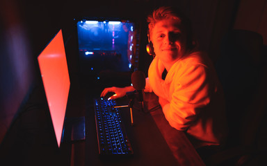 Wall Mural - Cute guy in headphones and casual clothes sits with microphone at a table and plays video games, looks into the camera with a smile.Portrait of a happy gamer at night at the computer,posing in camera