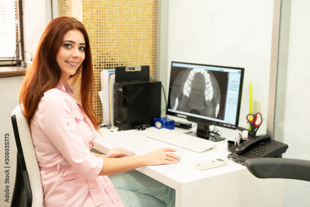 A female dentist doctor is sitting at a table, on a computer a CT scan of the jaw. The doctor is dressed in professional clothes. - obrazy, fototapety, plakaty 