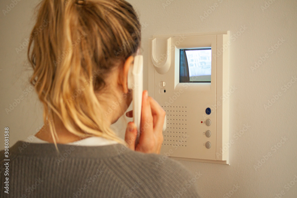 Blonde woman answering a call at a door phone while looking at the screen on the CRT display. Video intercom equipment. Selective focus image. - obrazy, fototapety, plakaty 