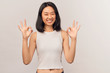 Girl shows two hands badge of okay, laughs, wrinkles nose from different jokes. Businesslike young woman Asian appearance dressed in short shirt stands isolated white background in Studio.