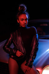 Wall Mural - a young beautiful afro-american girl in a leather jacket, in a bodysuit posing on the hood of a car. stylish street light. night.