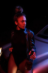 Wall Mural - a young afro-american beautiful  girl in a leather jacket,in a bodysuit and with a bottle of alcoholic beverage poses on the hood of a car. stylish street light. night.