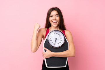 Pretty young girl with weighing machine over isolated pink background with weighing machine and doing victory gesture