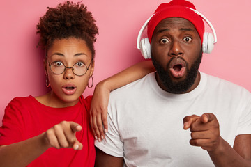 Wall Mural - Horizontal view of emotional stupefied ethnic young boyfriend and girlfriend point at camera, notice something unbelievable, have scared gazes, man wears stereo headphones on ears. Omg concept