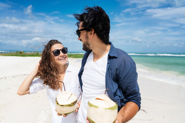 Wall Mural - Beautiful young couple joyfully on a tropical beach with coconuts in their hands on the seashore. Honeymoon Travel and Vacation on the Tropical Ocean