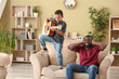 © Pixel-Shot - African-American man covering ears while his little son is playing guitar at home