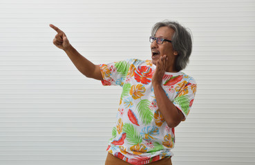 Excited elderly traveler asian man wearing summer shirt and glasses pointing finger away standing over white wall, Business summer holiday concept