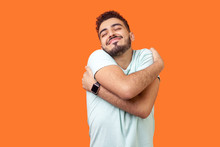 I Love Myself! Portrait Of Egoistic Brunette Man With Beard In White T-shirt Standing With Closed Eyes, Embracing Himself And Smiling Form Pleasure And Proud. Indoor, Isolated On Orange Background
