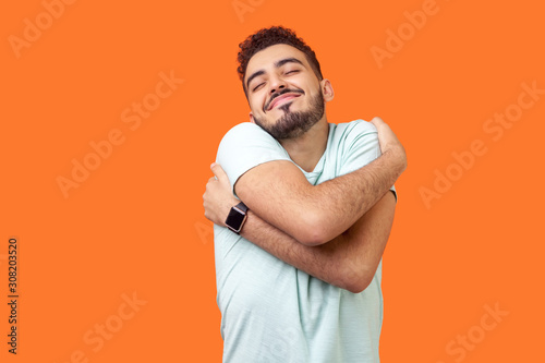 Cuadros en Lienzo I love myself! Portrait of egoistic brunette man with beard in white t-shirt standing with closed eyes, embracing himself and smiling form pleasure and proud