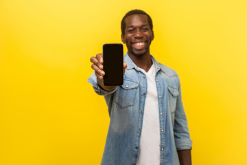 Online service, technology. Portrait of satisfied glad young man in denim casual shirt standing holding out cellphone and smiling broadly at camera. indoor studio shot isolated on yellow background