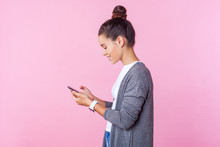 Side View Of Happy Beautiful Teenage Girl With Bun Hairstyle In Casual Clothes Using Cell Phone, Feeling Excited Of Chatting With Friends, Scrolling Social Network. Indoor Studio Shot, Pink Background